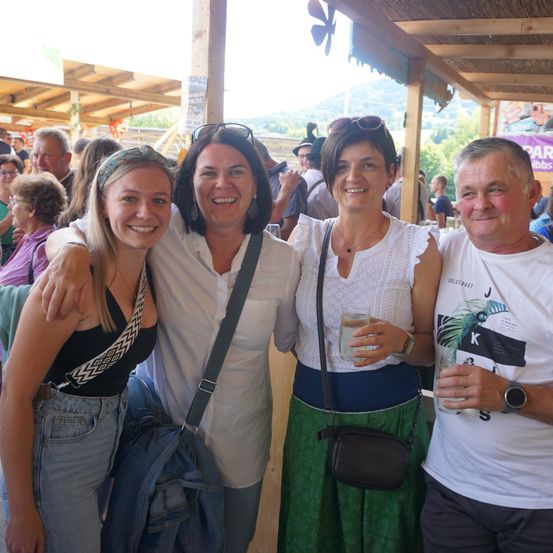 Vier Erwachsene, zwei Frauen und zwei Männer, stehen zusammen unter einem Pavillon bei einer Veranstaltung im Freien. Sie alle lächeln für die Kamera.