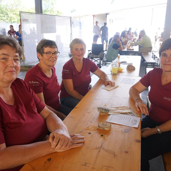 Vier Frauen in roten T-Shirts sitzen an einem Tisch in einem Essbereich. Dahinter sind Tische mit Essen, und Menschen sitzen. Die Wände sind weiß, und es gibt ein Schild mit Schrift.