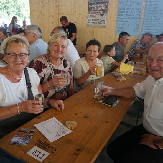 Mehrere Erwachsene sitzen an einem langen Holztisch, lächeln und halten Getränke. Sie befinden sich in einer rustikalen Umgebung mit Holzwänden. Im Hintergrund stehen Menschen und ein Plakat mit einem Datum.
