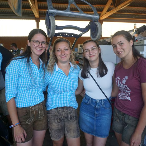 Vier Frauen in traditioneller Kleidung stehen zusammen unter einem Scheunendach. Die Frau auf der linken Seite trägt ein blau kariertes Hemd und braune Shorts. Die zweite Frau von links trägt ein blaues Hemd und braune Shorts. Die dritte Frau trägt ein weißes Hemd und eine blaue Jeansshorts. Die Frau auf der rechten Seite trägt ein lila Hemd.
