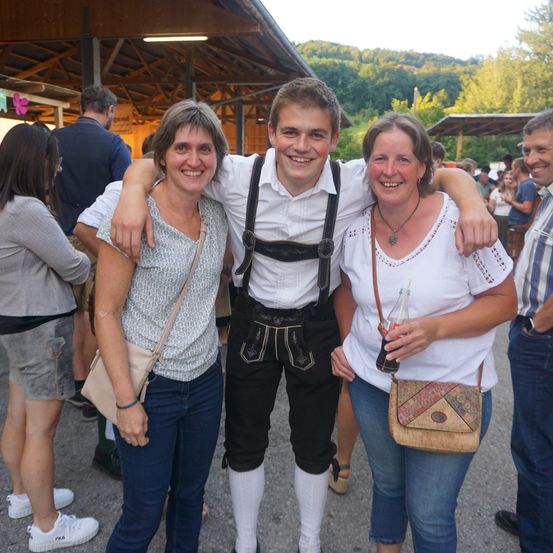 Ein junger Mann in traditioneller bayerischer Kleidung posiert mit zwei lächelnden Frauen, möglicherweise auf einem Festival. Hinter ihnen ist eine Menschenmenge, eine Struktur und eine Bergkulisse zu sehen.