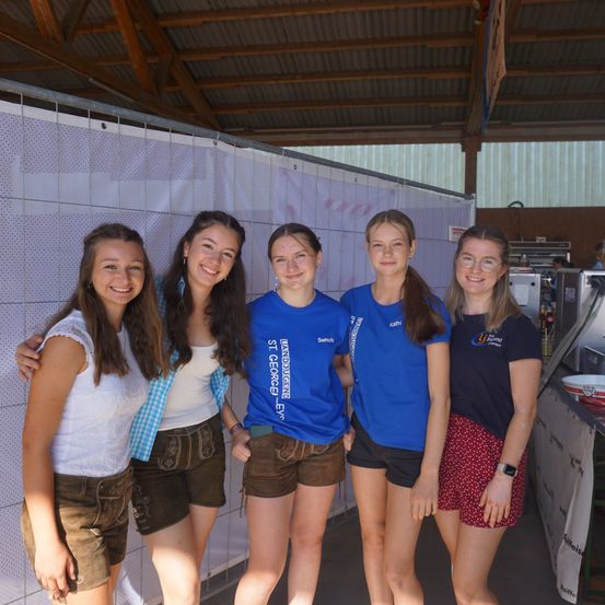 Fünf junge Frauen stehen in einem Raum und tragen traditionelle Kleidung. Drei von ihnen tragen blaue T-Shirts mit gedrucktem Text. Sie alle haben Lächeln auf ihren Gesichtern.