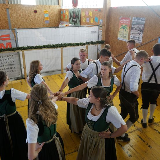 Mehrere Menschen in traditioneller Kleidung tanzen in einem Kreis in einem Raum mit Holzwänden und -boden. Einige Poster und Banner hängen an den Wänden.