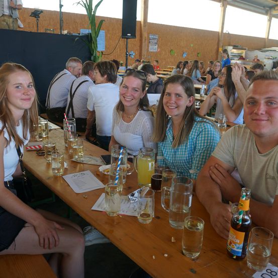 Eine Gruppe von Menschen, darunter vier Frauen und ein Mann, sitzen an einem langen Tisch mit Gläsern und Getränken. Sie scheinen eine gesellige Zusammenkunft zu genießen.