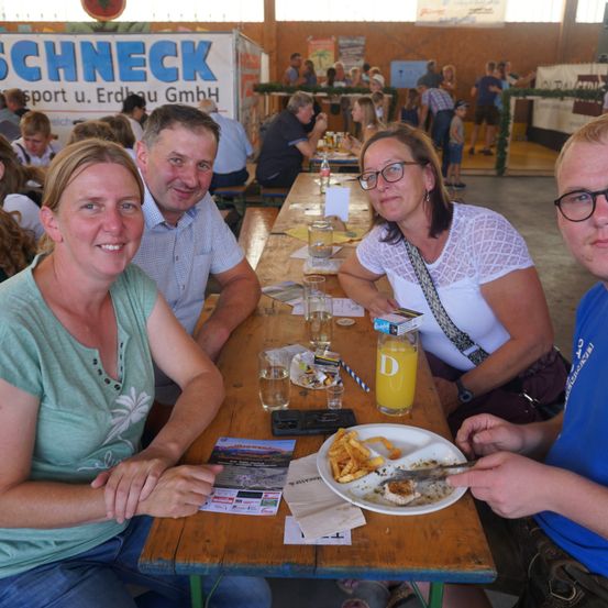 Vier Personen sitzen an einem Tisch in einer großen Halle. Sie haben Essen und Getränke vor sich. Die Frau auf der linken Seite lächelt in die Kamera. Der Mann neben ihr schaut in die Kamera. Die Frau auf der rechten Seite trägt eine Brille und schaut weg. Der Mann auf der rechten Seite trägt ein blaues Shirt.