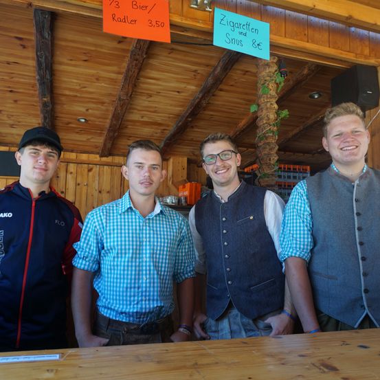 Vier Männer stehen vor einem Holztresen. Ein Mann trägt ein blau-weiß kariertes Hemd. Hinter ihnen hängen Schilder an der Decke.