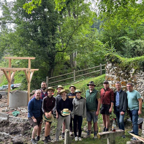 Eine Gruppe von Menschen posiert für ein Foto in einem Waldgebiet. Sie stehen auf dem Gras, und einige von ihnen tragen Hüte. Eine Holzstruktur ist auf der linken Seite platziert, und eine Steinmauer befindet sich auf der rechten Seite. Dahinter sind einige Bäume gepflanzt, und ein Holzzaun befindet sich am Hang.