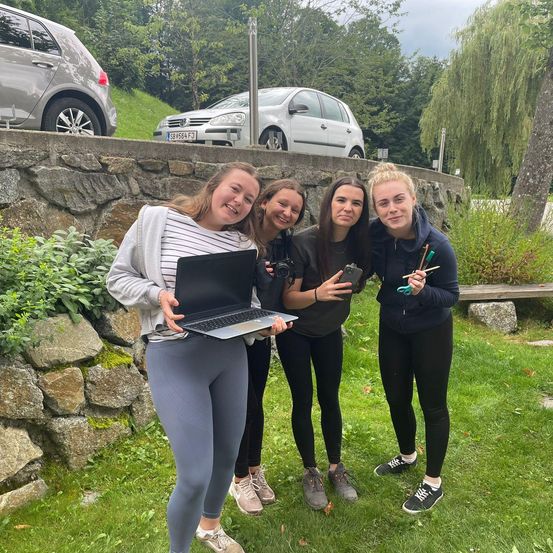 Vier Frauen posieren für ein Foto im Park. Eine hält einen Laptop, eine andere eine Kamera, während die anderen Pinsel halten. Hinter ihnen ist eine Steinmauer mit geparkten Autos.