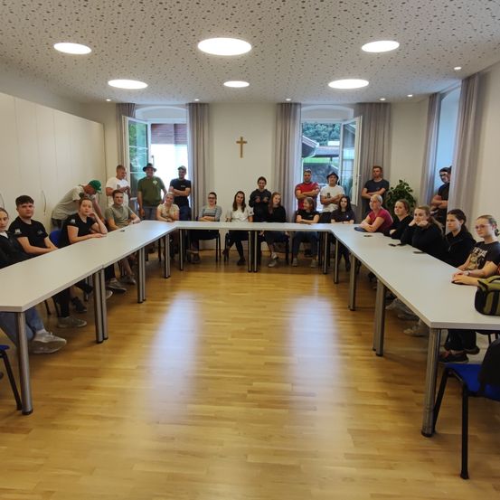 Eine Gruppe von Menschen sitzt um einen großen Tisch in einem Raum mit Holzboden und weißen Wänden. Einige tragen Freizeitkleidung, und an der Wand befindet sich ein Kreuz.
