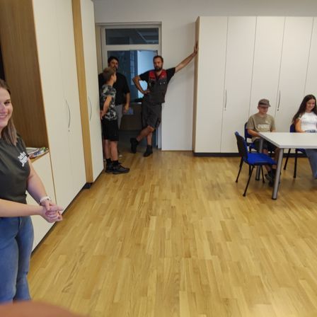 Mehrere Personen befinden sich in einem Raum mit Holzböden. Ein Mann steht mit der Hand an der Tür, während andere zuschauen. Eine Frau steht mit gefalteten Händen vor sich.