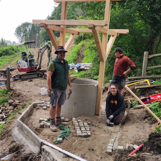 Drei Personen arbeiten auf einer Baustelle, stehend um eine Holzstruktur herum. Eine Person hockt in der Nähe eines Betonzylinders, während die anderen beiden stehen und sprechen. Ein Baufahrzeug ist hinter ihnen geparkt.
