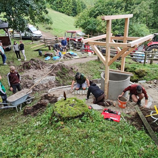 Eine Gruppe von Menschen arbeitet auf einer Baustelle. Sie benutzen eine Schubkarre, Schaufeln und andere Werkzeuge. Eine Holzkonstruktion ist in der Nähe, und einige Leute sind bei einem Betontank. Fahrzeuge und Bäume sind im Hintergrund.