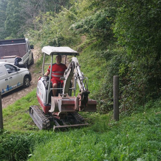 Ein Mann fährt einen rot-weißen Bagger auf einem steilen, grasbewachsenen Hang. Dahinter sind ein weißes Auto und ein Lastwagen auf der unbefestigten Straße geparkt.