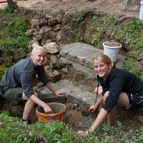 Zwei Frauen arbeiten auf einer Baustelle, eine gießt Zement in einen Eimer, und die andere sitzt neben ihr.