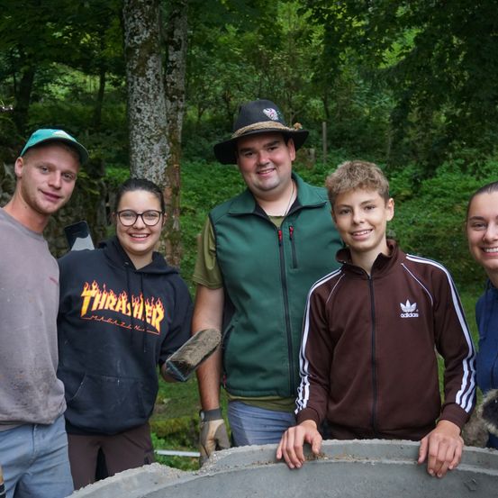 Fünf Menschen lächeln und posieren für ein Foto im Wald. Einer hält eine Kelle. Ein anderer trägt eine Thrasher-Hoodie. Ein Mann in einem grünen Vest steht hinter ihnen.