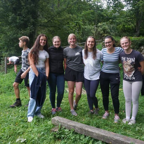 Eine Gruppe von sechs jungen Menschen steht auf einem Grasfeld und posiert für ein Foto. Bäume und ein Zaun sind hinter ihnen, mit einer Person, die sich bügt. Sie alle lächeln und scheinen sich zu amüsieren.