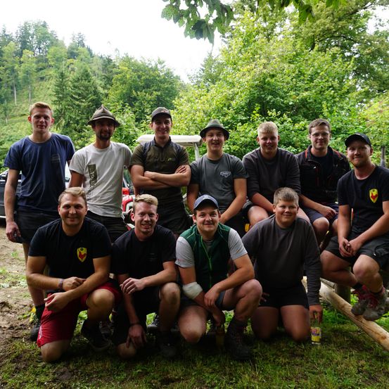 Eine Gruppe junger Männer posiert für ein Foto im Freien, sitzt auf Baumstämmen in einem Wald. Sie lächeln und tragen verschiedene Mützen und Freizeitkleidung. Im Hintergrund sind Bäume und ein Auto zu sehen.