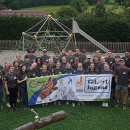 Eine Gruppe von Menschen in passenden grauen T-Shirts und mit einem Banner posiert für ein Foto vor einem Spielplatz.