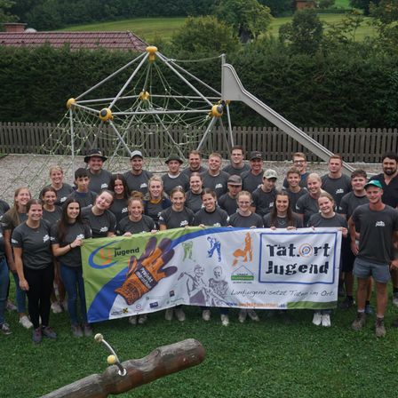 Eine Gruppe von Menschen in passenden grauen T-Shirts und mit einem Banner posiert für ein Foto vor einem Spielplatz.