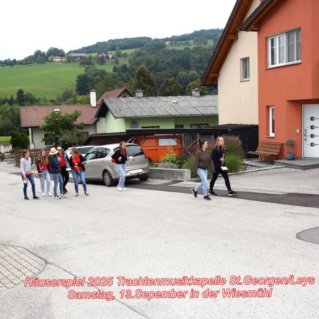Eine Gruppe von Menschen in roten Shirts und Hüten geht in einem Dorf, nahe Häusern und einem Auto, am 13. September 2025.