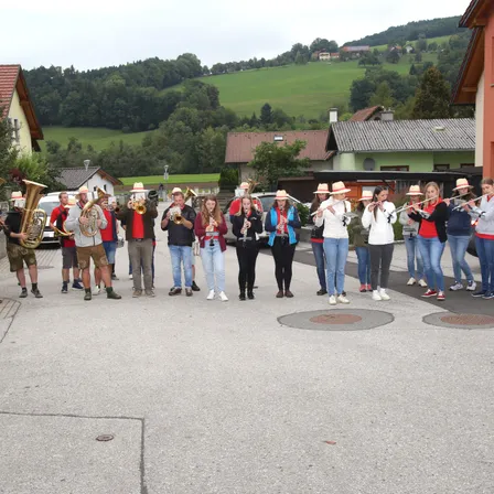 Eine Blaskapelle spielt auf der Straße, wobei die Musiker verschiedene Instrumente spielen. Im Hintergrund befinden sich Häuser und üppige grüne Hügel.