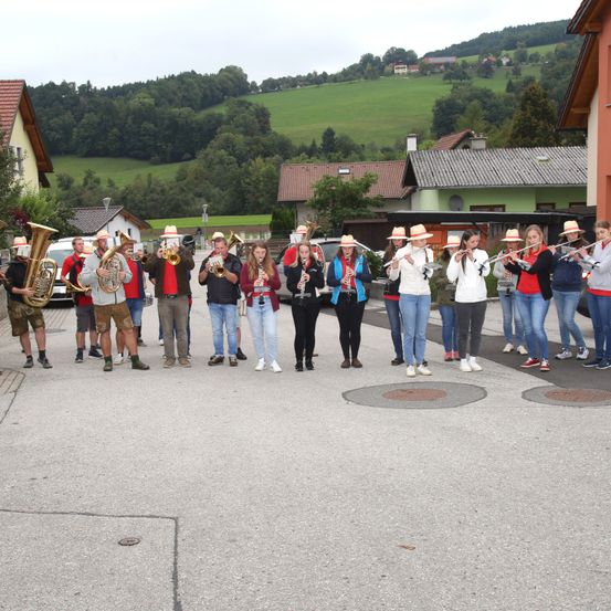Eine Blaskapelle spielt auf der Straße, wobei die Musiker verschiedene Instrumente spielen. Im Hintergrund befinden sich Häuser und üppige grüne Hügel.