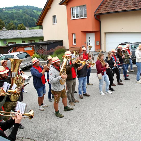 Eine Blaskapelle in Hüten und Uniformen steht vor einem Haus und tritt auf. Die Band besteht aus Tuben, Trompeten und anderen Instrumenten.