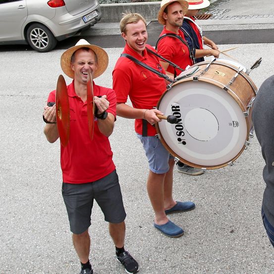 Drei Männer in roten Hemden und Hüten spielen Instrumente im Freien. Ein Mann hält eine große Trommel, ein anderer spielt ein Metallinstrument und ein dritter ein Drumstick.