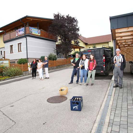 Eine Gruppe von Menschen steht vor einem Gebäude. Ein Mann mit Hut hält eine Flasche und eine Frau eine Tasche. In der Nähe befinden sich Bierkisten und ein Lieferwagen.