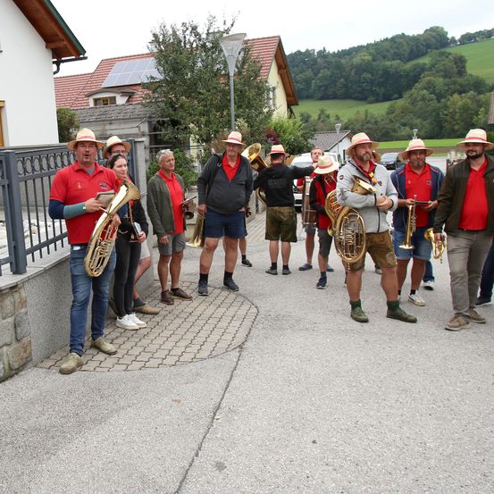 Eine Blaskapelle in roten Oberteilen posiert für ein Foto im Freien, auf einem Kopfsteinpflasterweg in einem Dorf mit Häusern, Grünflächen und Hügeln im Hintergrund.