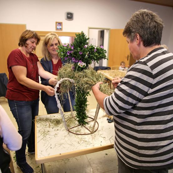 Drei Frauen fertigen eine Blumenarrangement auf einem Tisch in einem Raum an. Eine hält einen Kranz, während eine andere Blumen arrangiert. Ein Baby sitzt in der Nähe.