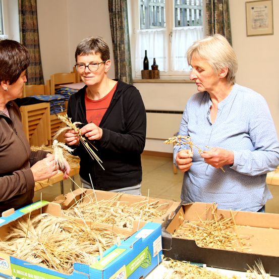 Drei Frauen stehen um einen Tisch mit Weizenähren herum. Sie halten und untersuchen den Weizen. Der Raum hat ein Fenster mit weißen Vorhängen.