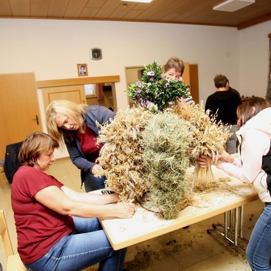 Eine Gruppe von Frauen in einem Raum kreiert eine Blumenarrangement aus Weizen und anderen Blumen auf einem Tisch.