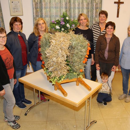 Eine Gruppe von Erwachsenen und Kindern steht um einen Tisch mit einer großen Blumenarrangement. Die Kinder hocken sich hin, und eines hält eine Pflanze. Dahinter steht ein Kreuz und eine weiße Wand.