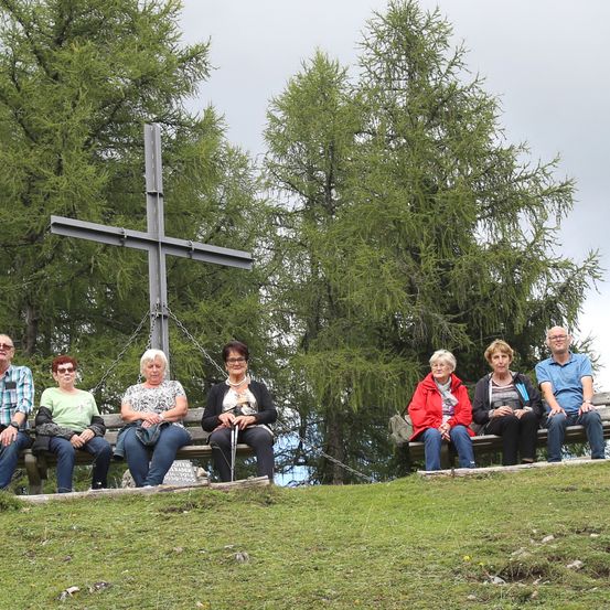 Sechs ältere Personen sitzen auf Bänken vor einem großen Kreuz, vor dem Hintergrund von immergrünen Bäumen und einem grasigen Hügel.