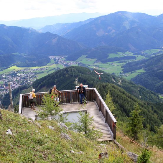 Drei Personen stehen auf einer hölzernen Aussichtsplattform hoch auf einem Berg und blicken auf ein Tal mit einer Stadt und grünen Feldern, mit entfernten Bergen im Hintergrund.
