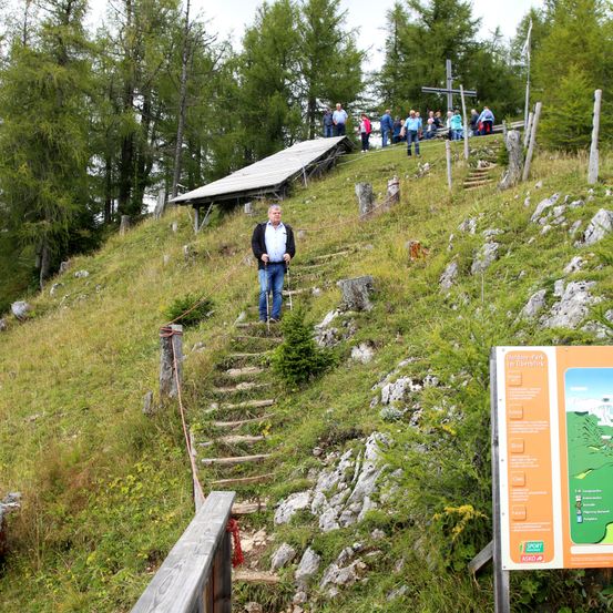 Ein Mann geht einen grasigen Hügel mit Holzstufen hinauf. Ein Solarpanel ist in der Nähe. Leute stehen oben, einige mit Rucksäcken.