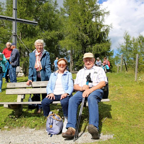 Eine Outdoor-Szene mit einem älteren Paar, das auf einer Bank sitzt, umgeben von anderen, die stehen und sitzen. Das Paar lächelt und scheint seinen Tag zu genießen. Hohe Bäume und ein großes Kreuz sind im Hintergrund.