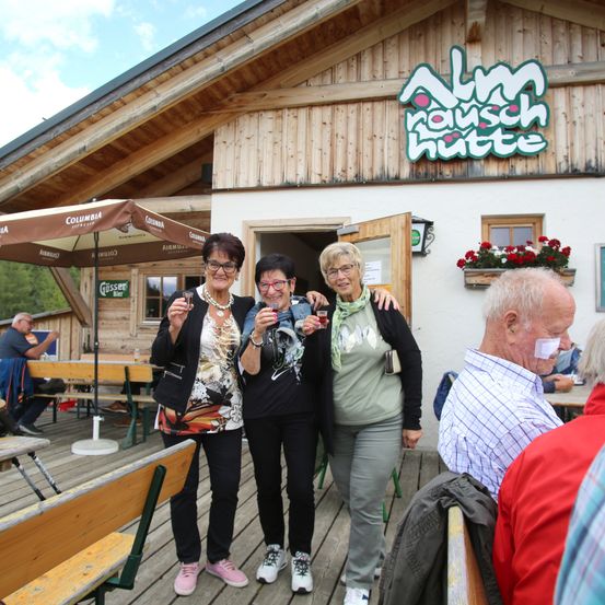 Drei Frauen stehen zusammen auf einer Holzterrasse, halten Getränke in den Händen und posieren für ein Foto. Hinter ihnen befindet sich ein Gebäude mit einem Schild, auf dem Alm Rausch Hut steht. Ein Mann sitzt im Hintergrund.