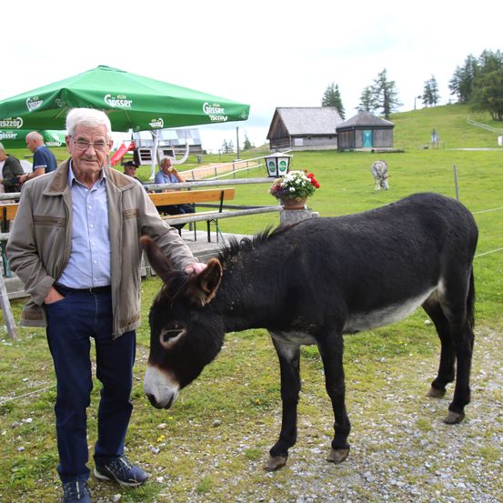 Ein älterer Mann steht neben einem schwarzen Esel auf einem Grasfeld. Hinter ihm ist ein grüner Schirm aufgestellt. In der Ferne befinden sich Gebäude und einige Menschen.