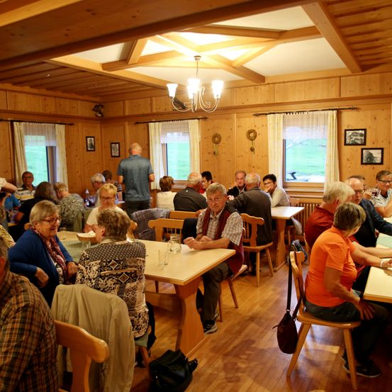 Eine Gruppe von Menschen diniert in einem gemütlichen Holzrestaurant mit Holzboden, -wänden und -decke. Sie sitzen um Tische herum, einige stehend, unterhalten sich und genießen ihre Mahlzeiten.