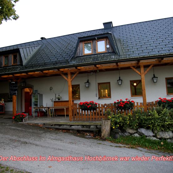 Ein weißes Gebäude mit grauem Dach und Holzveranda. Es hat mehrere Fenster und rote Blumen am Zaun. Der Text am unteren Rand lautet 'Der Abschluss im Almgasthaus Hochbarnek war wieder Perfekt!'.