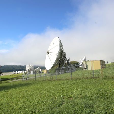 Ein Feld mit mehreren Satellitenschüsseln und einem Maschendrahtzaun unter einem teilweise bewölkten Himmel.