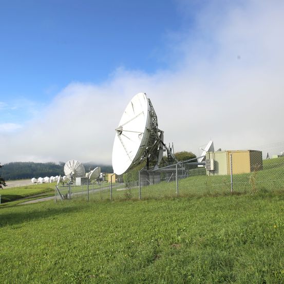 Ein Feld mit mehreren Satellitenschüsseln und einem Maschendrahtzaun unter einem teilweise bewölkten Himmel.