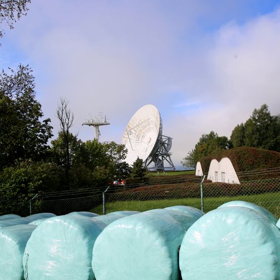 Eine Landschaft mit mehreren großen, zylindrischen Objekten in Plastik eingewickelt und einer großen Satellitenschüssel auf einem Hügel unter einem bewölkten Himmel.