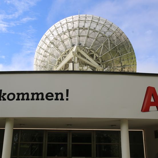 Ein Gebäude mit einer großen Satellitenschüssel auf der Spitze, die Worte 'kommen!' sind auf der Seite geschrieben. Das Gebäude hat Glastüren und Fenster.