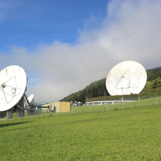 Mehrere Satellitenschüsseln stehen auf einer grünen Wiese, mit einem Gebäude und Bäumen im Hintergrund. Der Himmel ist blau mit verstreuten Wolken.