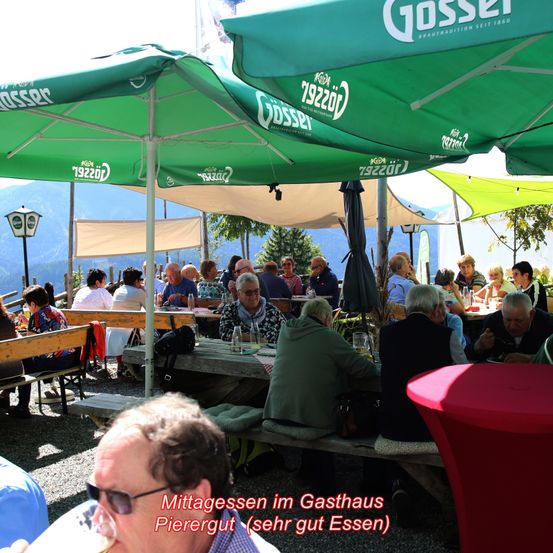 Mehrere Personen sitzen unter grünen Sonnenschirmen an Tischen in einem Gasthaus. Ein Mann mit Sonnenbrille trinkt aus einem Glas. Der Textüberlagerung lautet Mittagessen im Gasthaus Pierergut (sehr gut Essen).