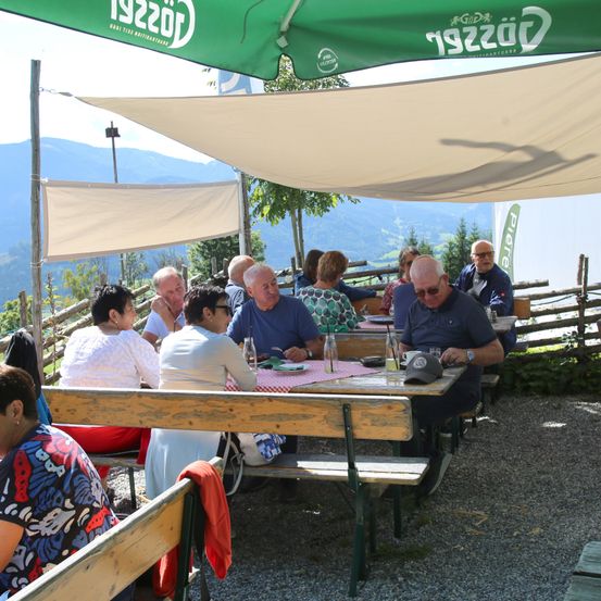 Eine Gruppe von Menschen sitzt unter einem grünen Sonnendach in einem Outdoor-Essbereich um einen Tisch herum, mit einer malerischen Bergsicht im Hintergrund.