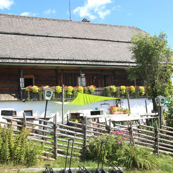 Ein traditionelles Holzhaus mit einem grünen Vordach, farbenfrohen Blumenkästen und einem eingezäunten Essbereich im Freien in einer sonnigen ländlichen Umgebung.